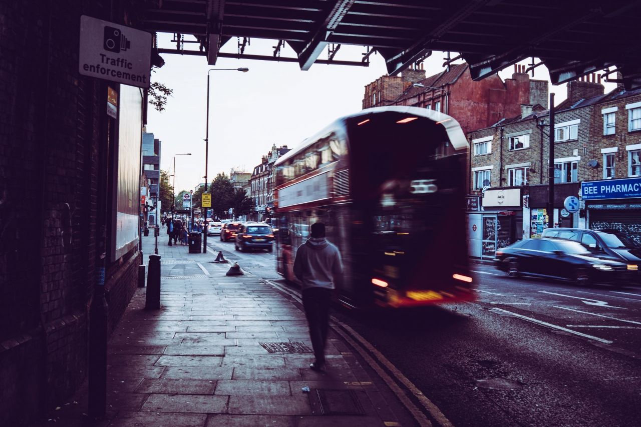 A photograph of an urban street. Featuring a pedestrian on a path and a bus on the road next to them. As a recent report highlighted concerns for vulnerable road users.