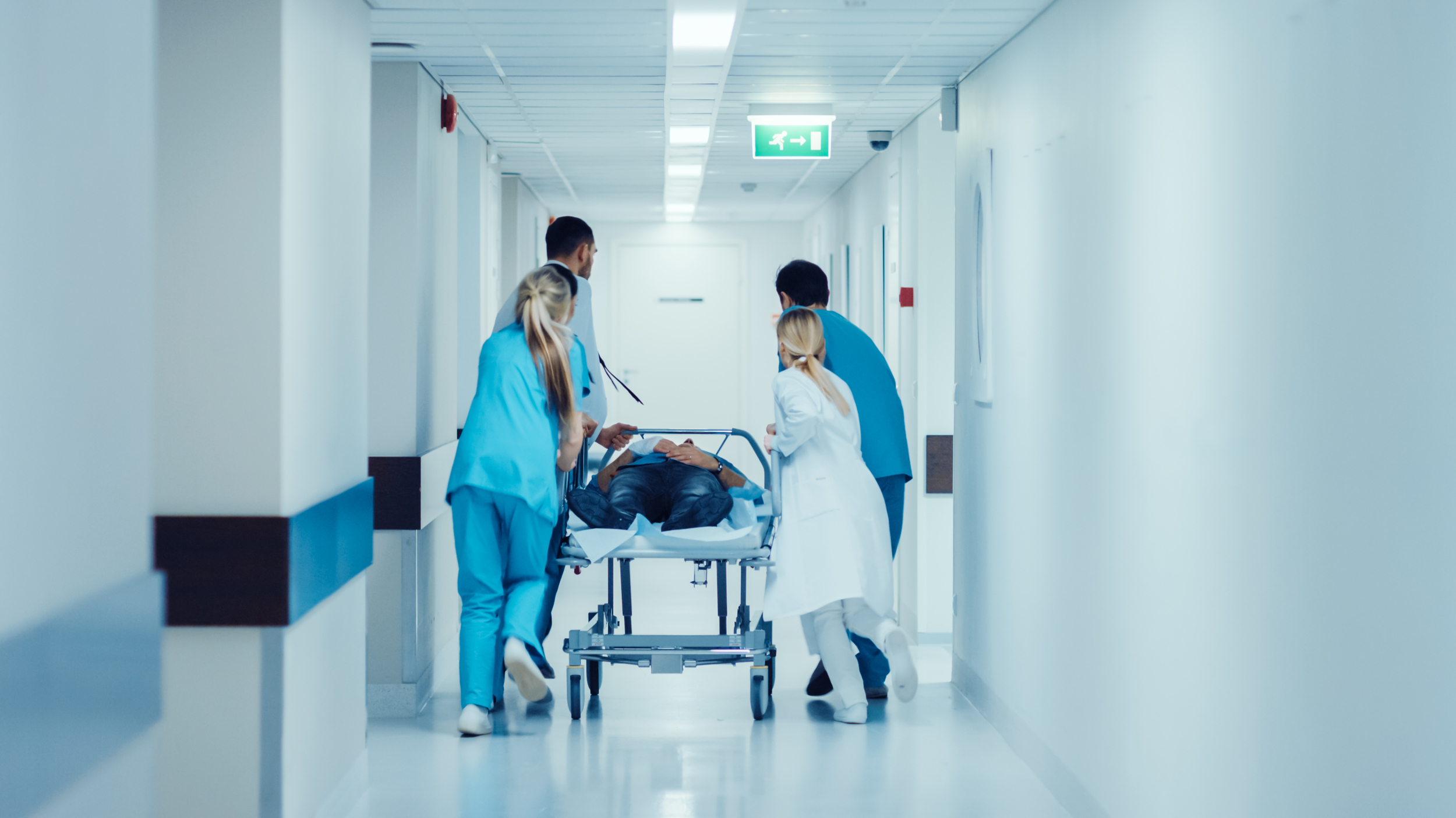 A photograph of a patient being wheeled through a hospital, on a bed, by medical staff.