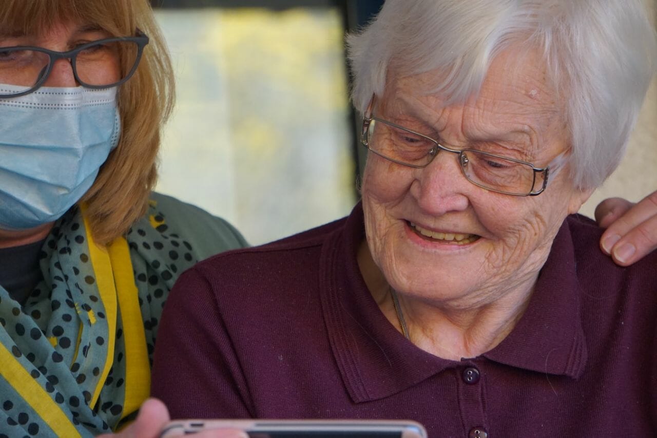 A photograph of an elderly lady, with somebody next to her holding a phone, as she smiles. The English government has been described as having a negligent approach to social care, in a damning report by the public accounts committee.