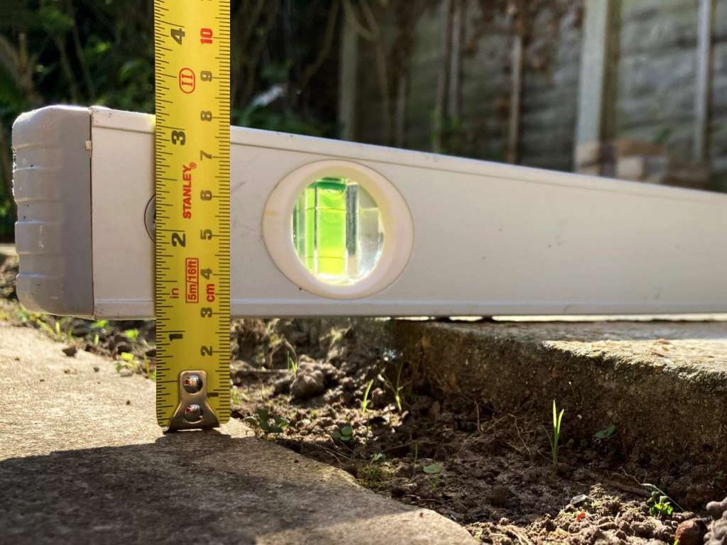 A photograph of a raised paving slab being measured. The same technique would be used when taking a photo of a pothole. It shows a straight edge over the top of the raised edge and a tape measure from the straight edge to the ground.